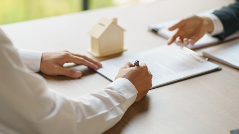 Osoba potpisuje ugovor za stolom dok na stolu stoji mala maketa kuće kao simbol zakupa nekretnine