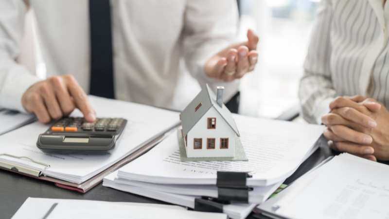 Knjigovođa sa kalkulatorom objašnjava finansijske obaveze dok maketa kuće stoji na dokumentima o zakupu