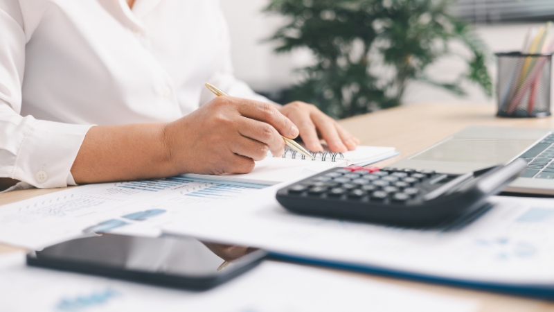 Osoba zapisuje podatke uz kalkulator i papire za obračun doprinosa na radnom stolu