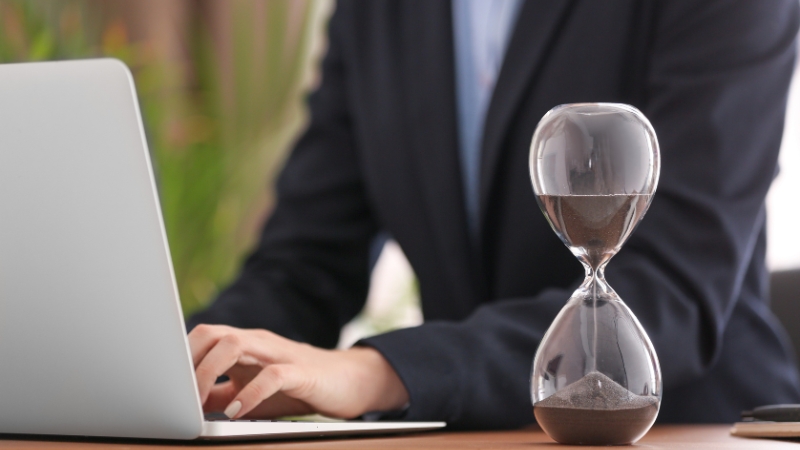 Osoba radi na laptopu pored peščanog sata koji simbolizuje prijavu na pola radnog vremena