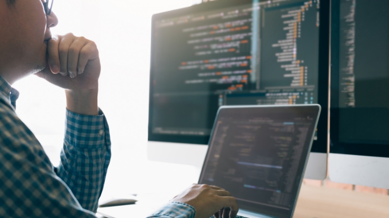 Programer radi za računarom i analizira kod na više ekrana, što jasno prikazuje šifra delatnosti 6201 kroz razvoj i obradu softvera