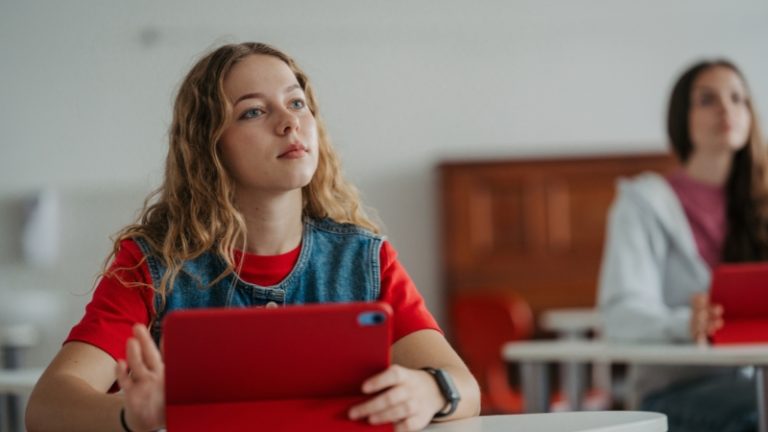 Devojka sedi u učionici i drži tablet, pažljivo prati nastavu dok druga učenica sedi iza nje