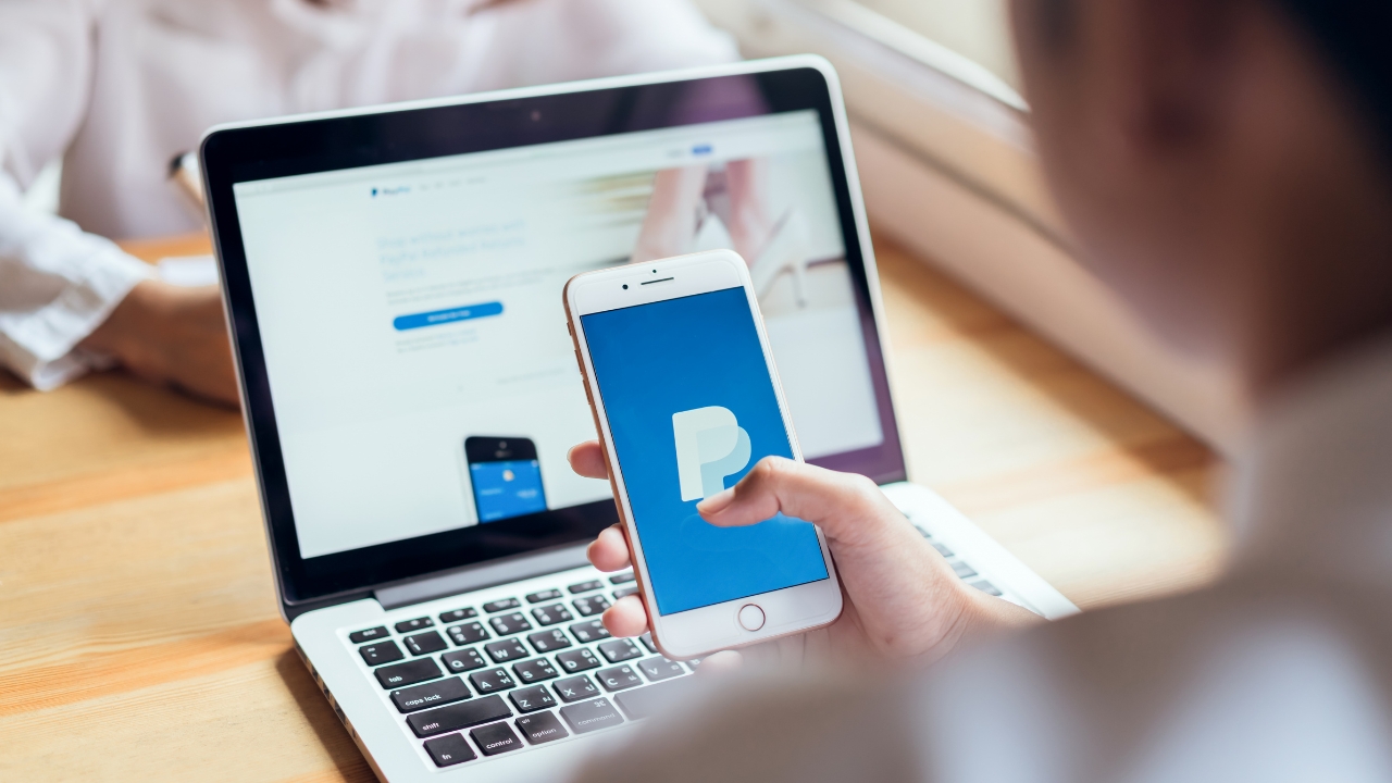 Osoba drži telefon sa PayPal aplikacijom ispred laptopa na kojem je otvoren PayPal sajt