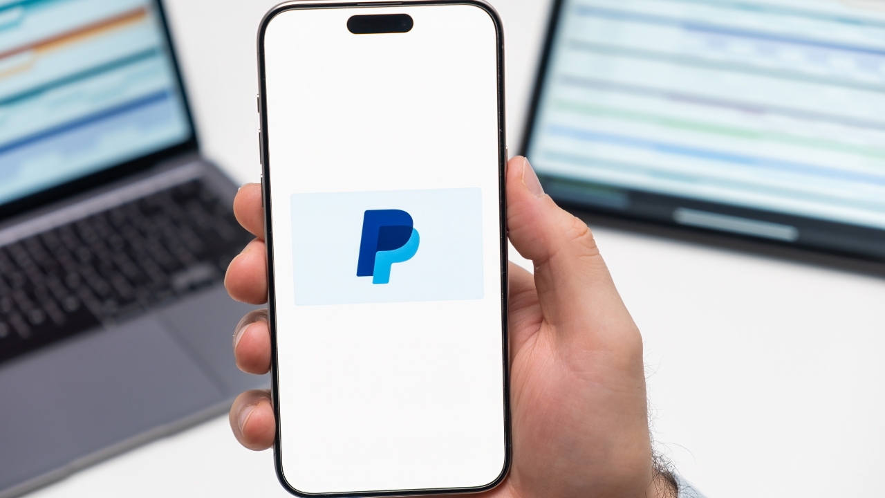 Osoba drži telefon sa PayPal logotipom ispred laptopa na stolu